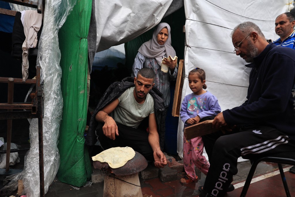 An internally displaced Palestinian man living in a shelter set up in a school run by the UN agency for Palestinian refugees (UNRWA), cooks a piece of unleavened bread on an open fire in a wet courtyard following overnight rainstorms in Rafah in the southern Gaza Strip on November 15, 2023