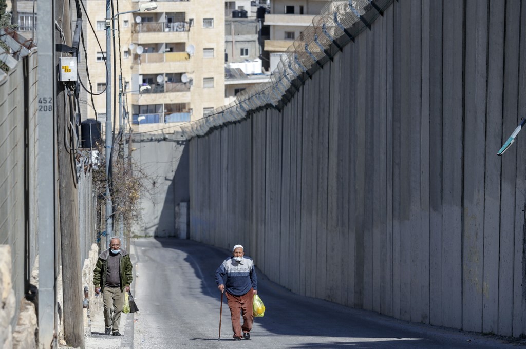 Palestinians walk by Israel’s controversial wall separating the occupied West Bank city of Abu Dis and East Jerusalem on 16 February 2021 (AFP)